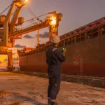 Робітник на причалі оглядає вантажне судно під вечірнім небом, демонструючи промислову діяльність.
