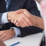 Два бізнесмени тиснуть руки через стіл у офісній обстановці, символізуючи угоду та партнерство.