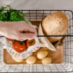 Рука тримає кошик з хлібом, картоплею, помідорами та зеленню, з верхнього ракурсу.