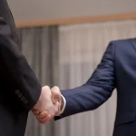 Двоє чоловіків у ділових костюмах тиснуть руки під час зустрічі.