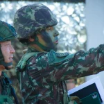 Група військовослужбовців у камуфляжі та шоломах обговорює тактичний план на відкритому повітрі.